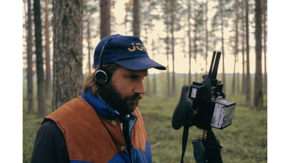 En man står i skogen med hörlurar och tittar in i en kamera