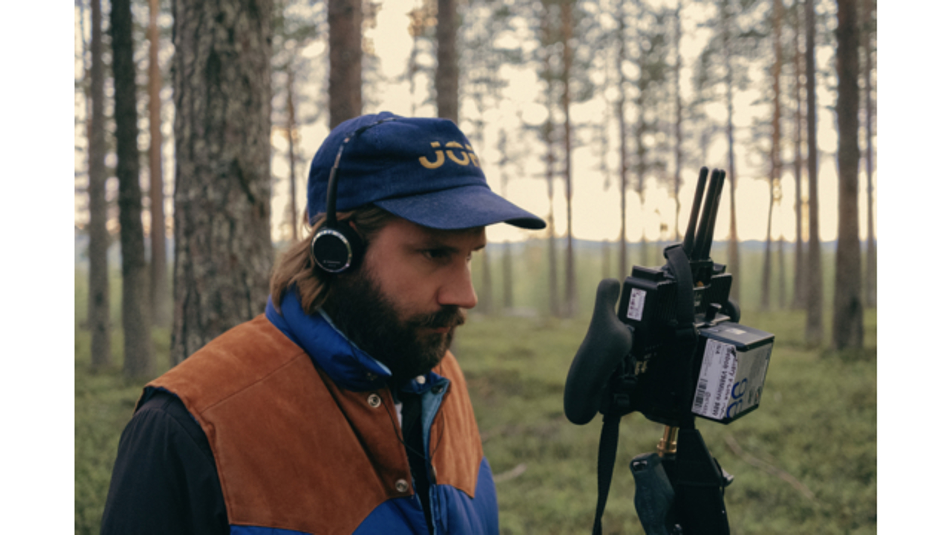 En man står i skogen med hörlurar och tittar in i en kamera