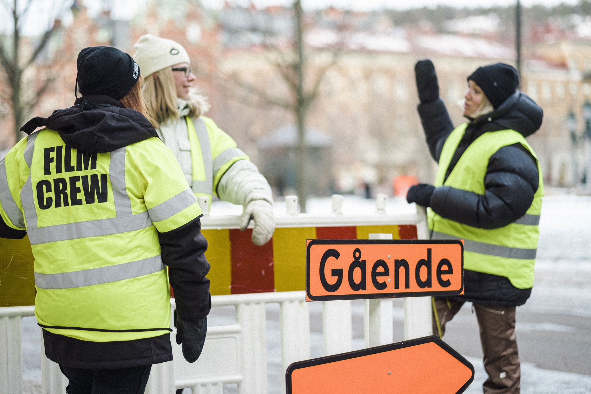 Tre människor med gula västar står utomhus och bevakar ett filminspelningsområde.
